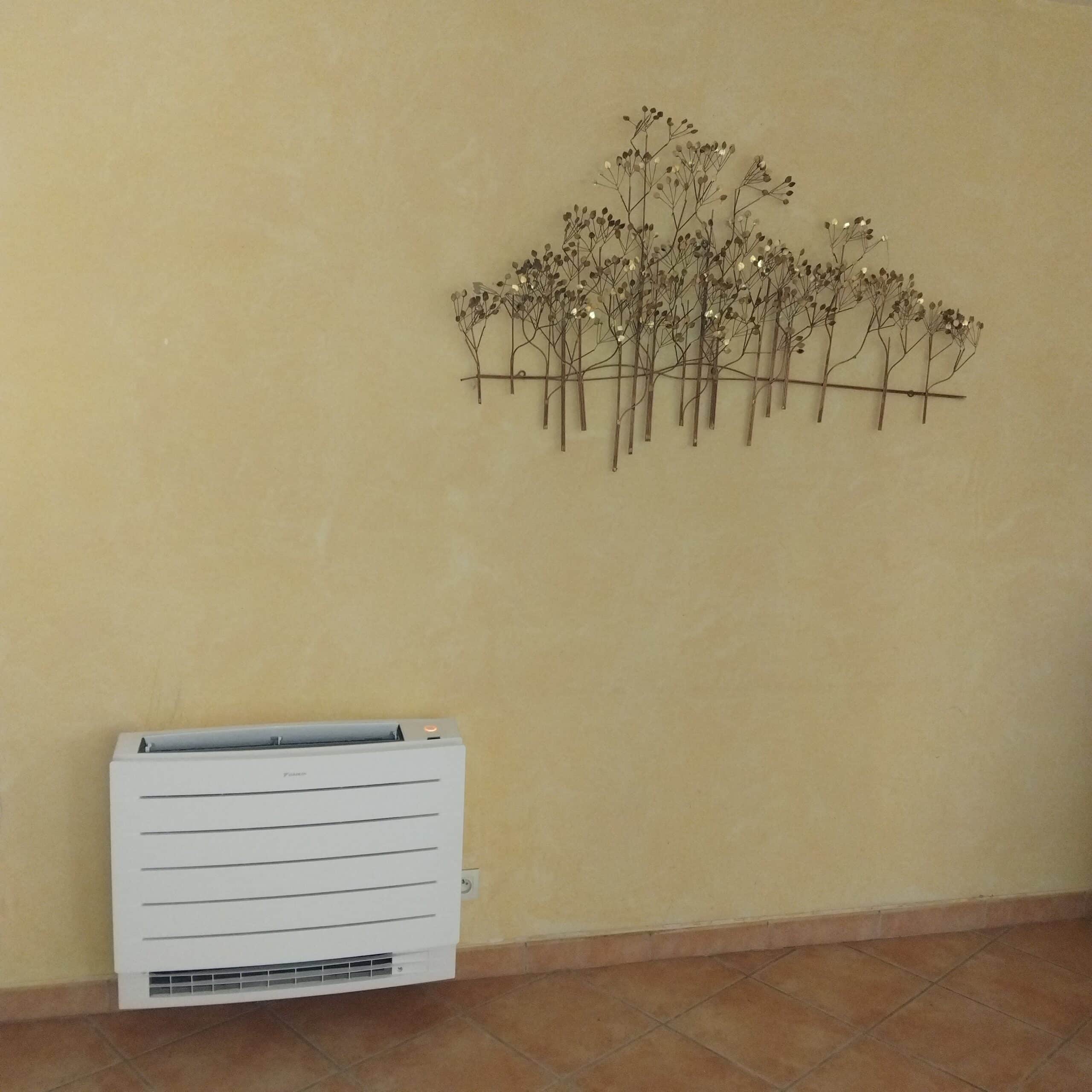 Climatiseur mural Daikin blanc sous une décoration murale en métal d'arbres sur un mur jaune texturé et sol carrelé.