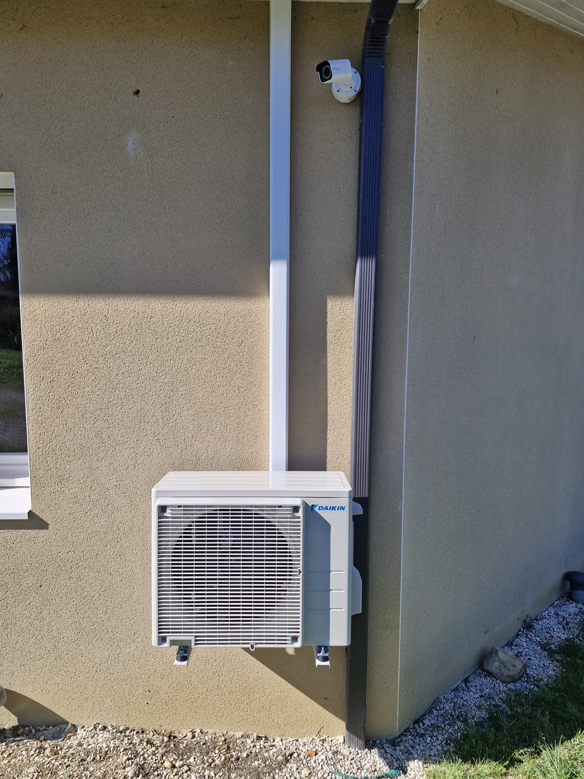 Unité extérieure Daikin installée sur un mur beige avec goulotte blanche, proche d'une descente de gouttière et caméra.