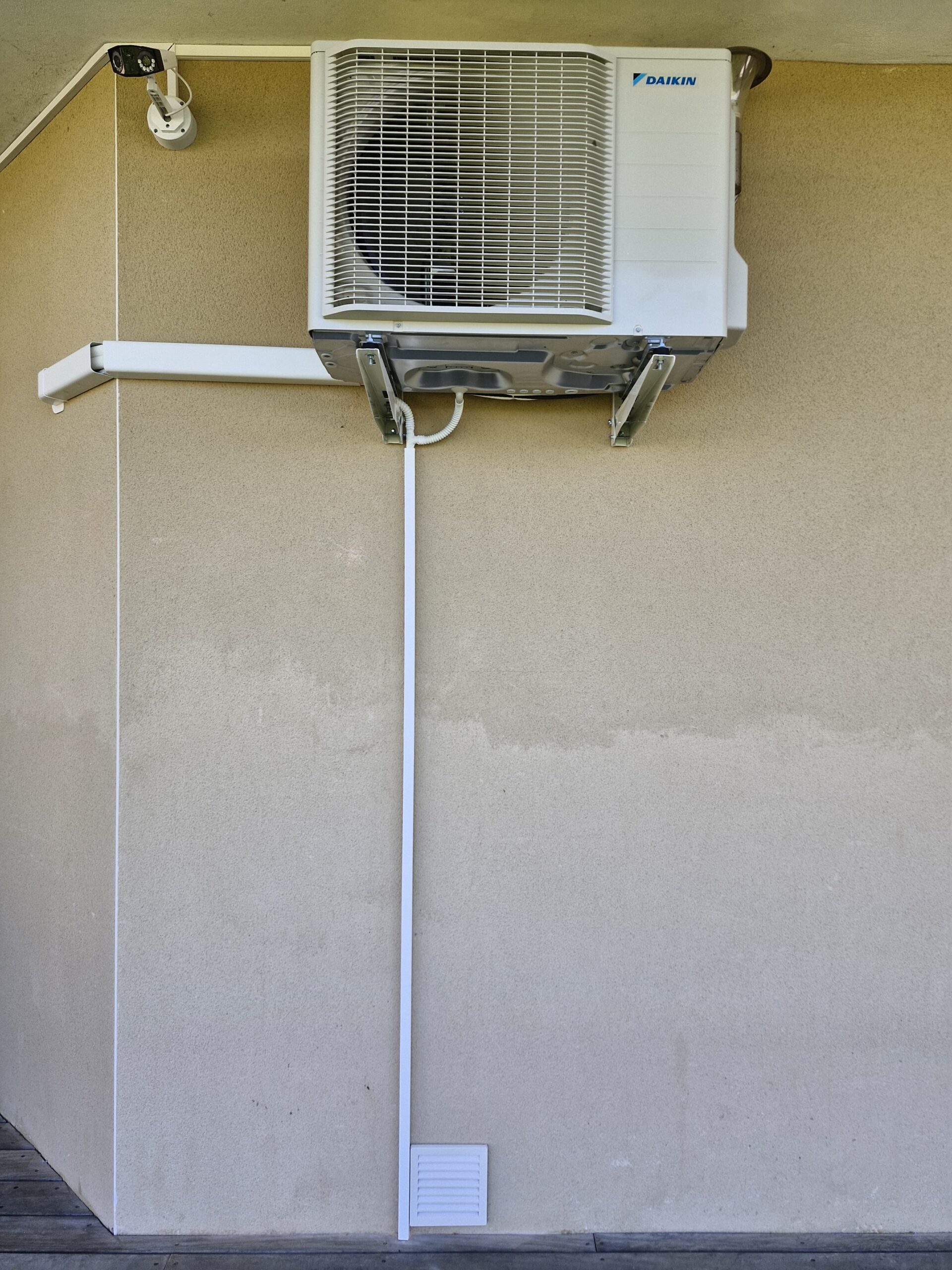 Unité extérieure Daikin montée sur mur beige avec conduit blanc, caméra de sécurité et grille de ventilation.