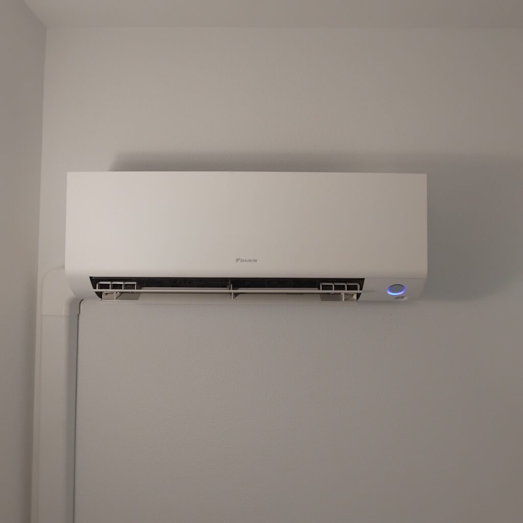 Climatiseur Daikin Mural Moderne : Performance et Design Climatiseur mural Daikin blanc allumé avec voyant bleu sur un mur gris, volets d'aération ouverts.