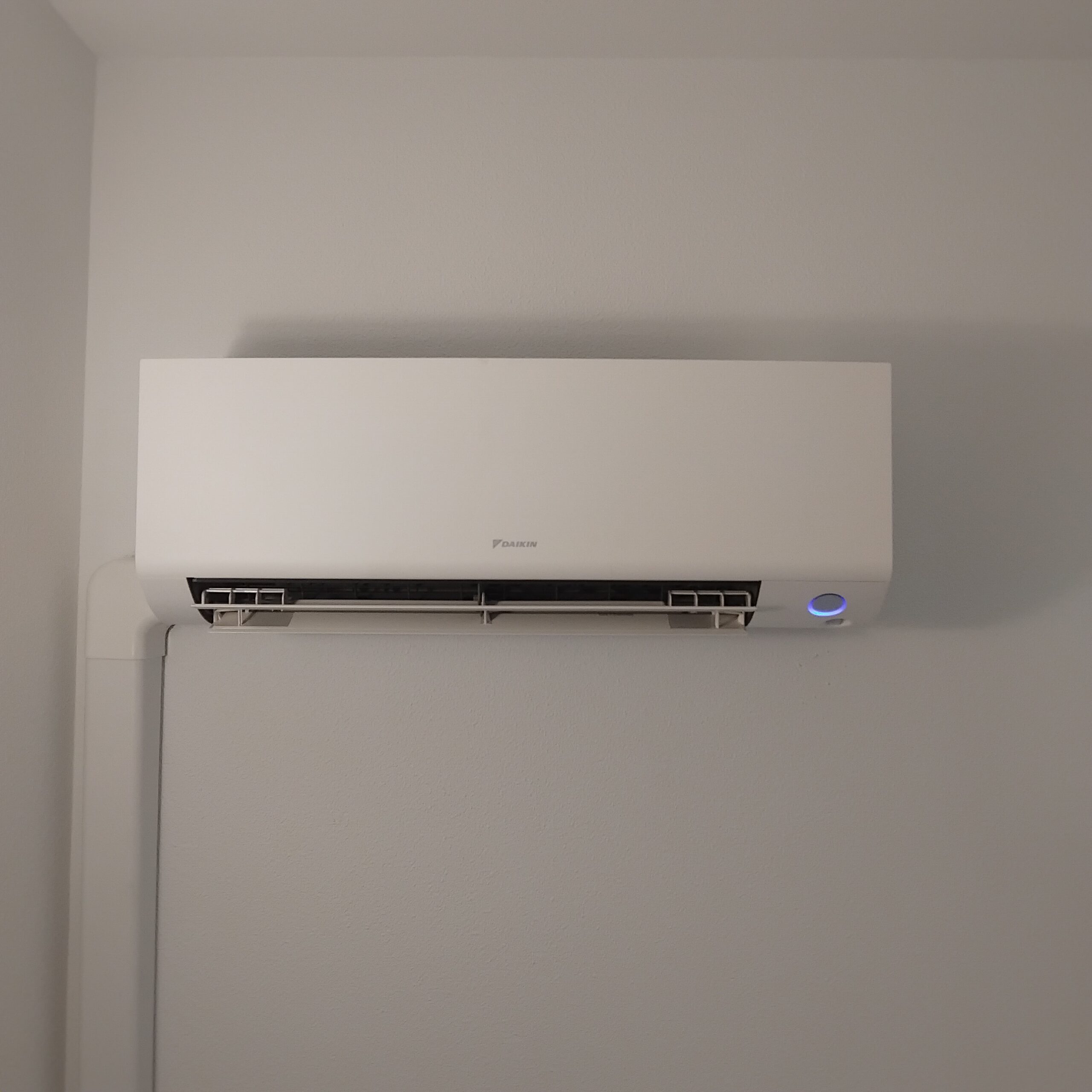 Climatiseur mural Daikin blanc allumé avec voyant bleu sur un mur gris, volets d'aération ouverts.