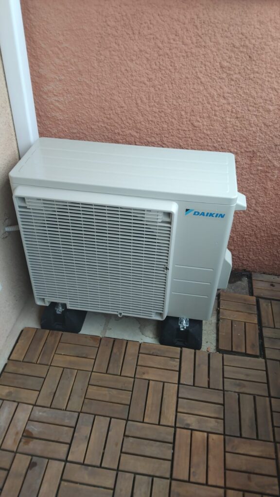 Climatisation Daikin extérieure : installation sur terrasse Climatiseur extérieur Daikin sur terrasse en bois contre un mur rose.