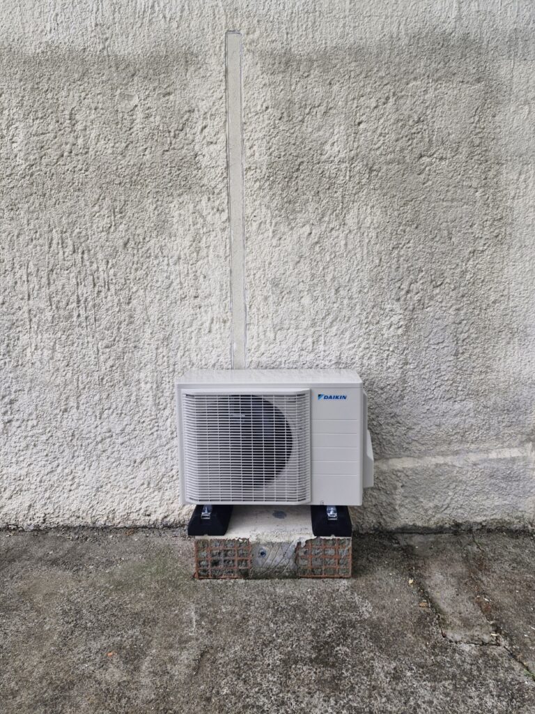 Installation d'un climatiseur Daikin extérieur mural Unité extérieure de climatisation Daikin installée sur un socle devant un mur texturé avec conduit vertical.